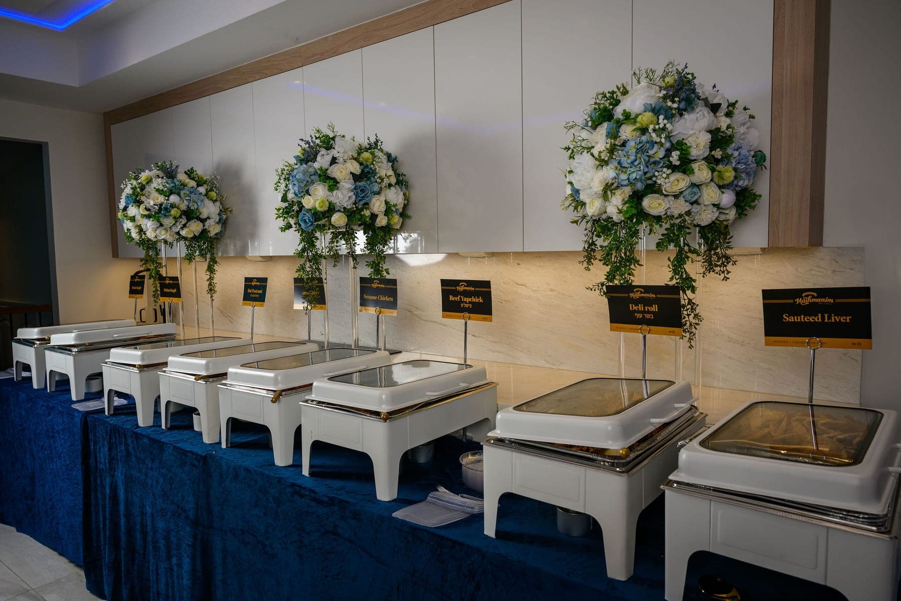 The buffet line receding down the room, white chafing dishes on blue-skirted tables under floral arrangements