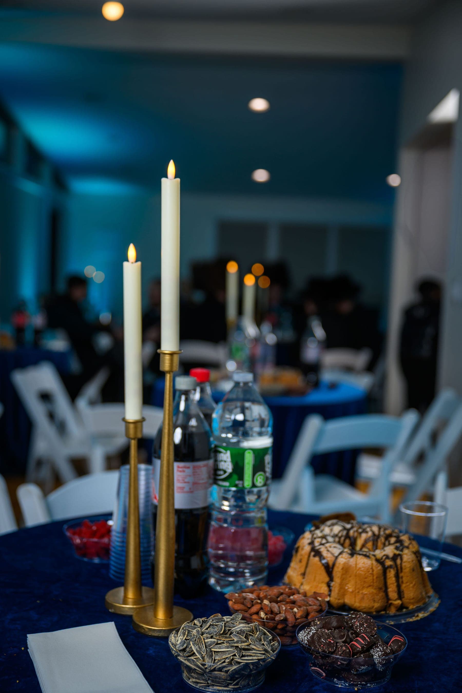 Two lit tapers in brass candlesticks on a round table dressed in deep blue, the room behind them out of focus