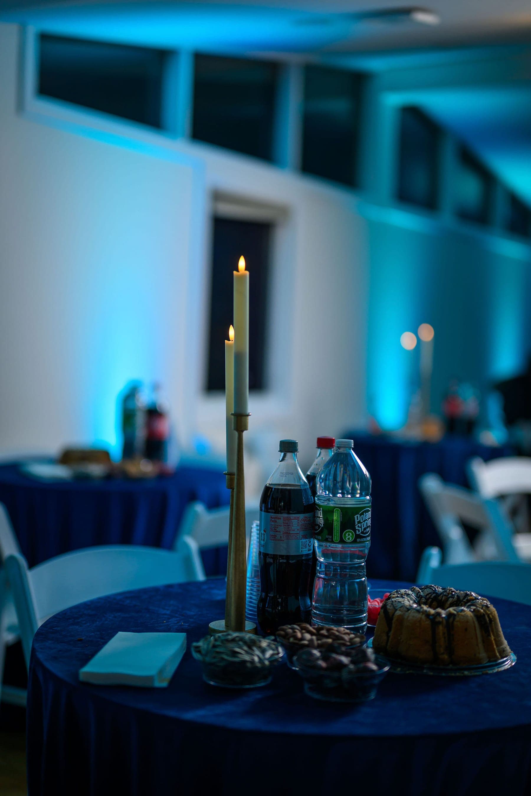 Round tables in blue velvet under blue uplighting, each with candles and a cake